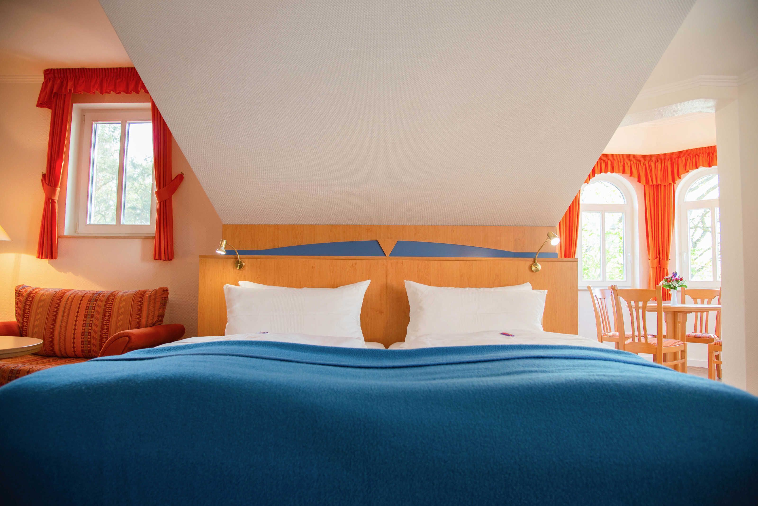 A hotel room with a large bed, a wooden headboard, a dining table with chairs, and two windows with red curtains.
