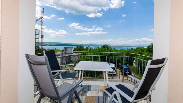 A balcony with a table and chairs, a view of the sea, and a construction crane in the background.