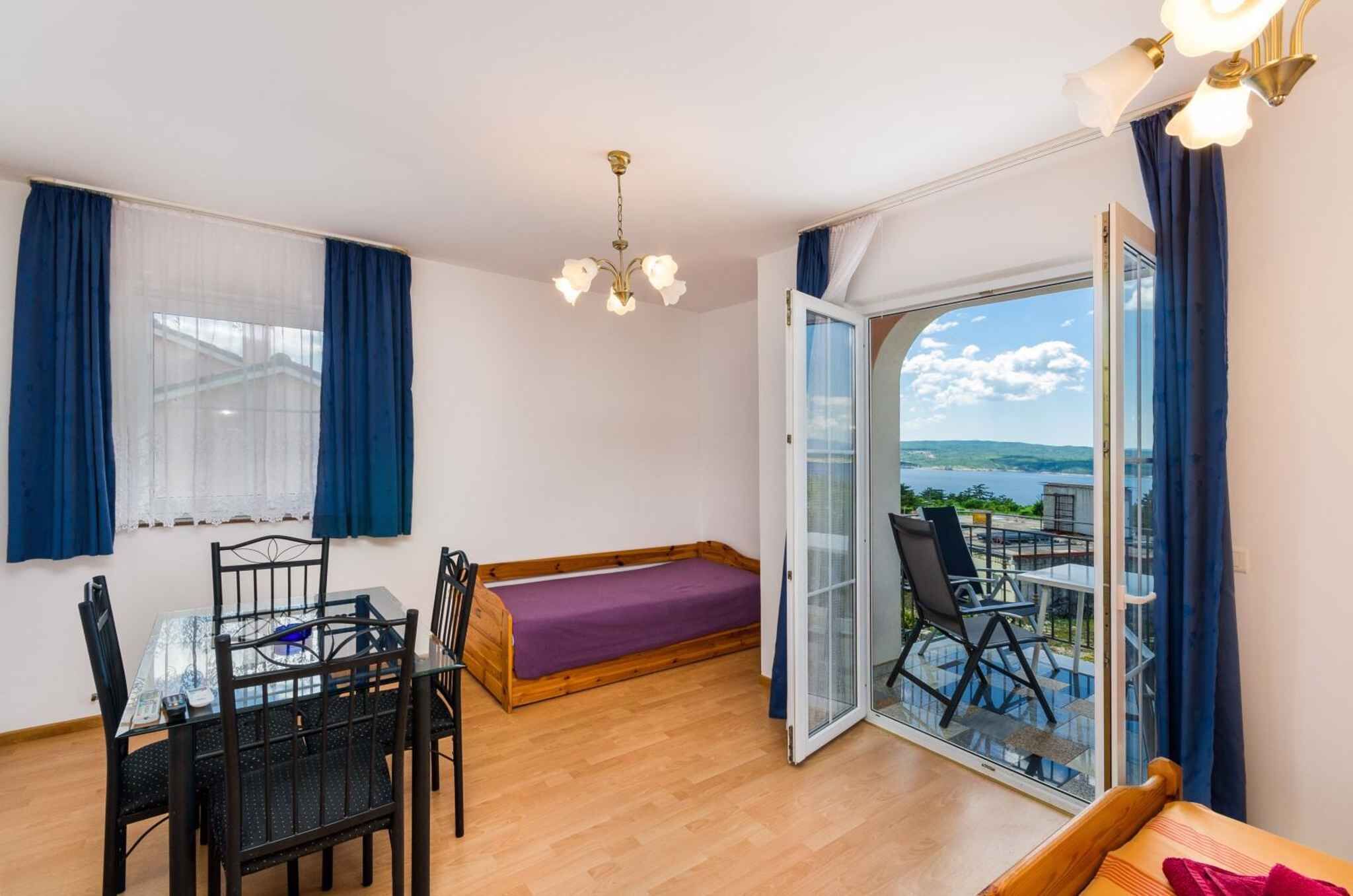 A room with a bed, a dining table with chairs, a chandelier, and a balcony with a view.