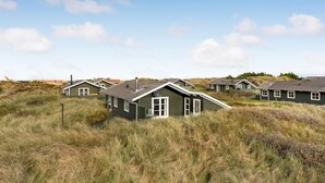 En række huse med grønne tage og hvide detaljer, beliggende i et græslandskab med klitter i baggrunden.