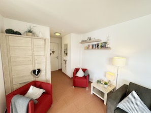 A living room with a red sofa, a grey sofa, a small table with a lamp, and a bookshelf with books and decorative items.
