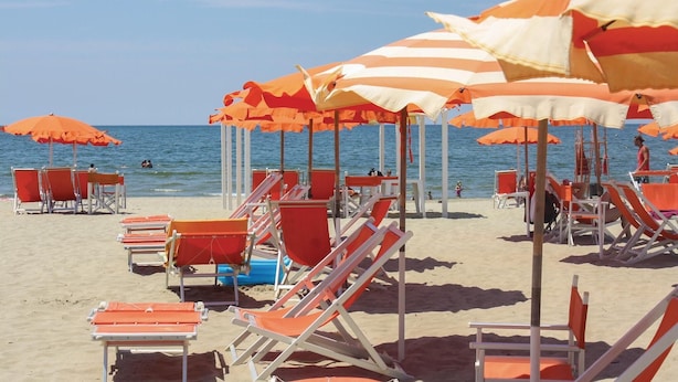 Ein Strand mit Reihen von orange-weiß gestreiften Sonnenschirmen und roten und weißen Liegestühlen.
