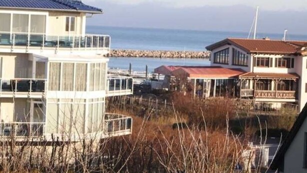 A coastal building with a balcony overlooking the sea, a pier in the background, and a small structure with a red roof. - (274) 1-room apartment - apartment complex Yachthafenresidenz (Kühlungsborn)