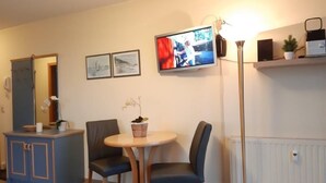 A hotel room with a bed, a desk with a chair, a small round table with two chairs, a television mounted on the wall, a floor lamp, and a shelf with a plant and a speaker. - (274) 1-room apartment - apartment complex Yachthafenresidenz (Kühlungsborn)