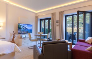 A hotel room with a bed, a TV, a dining table with chairs, a sofa, and a balcony with a view of trees.