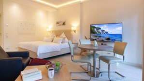 Une chambre d’hôtel comprenant un lit, une table à manger avec des chaises, un téléviseur et un tableau représentant un paysage côtier.