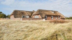 Une maison au toit de chaume, aux murs en briques, entourée d’herbes hautes.