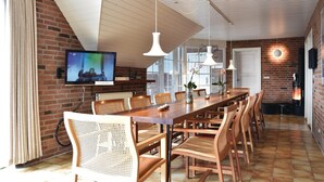 Un espace repas moderne avec une longue table en bois, des chaises en osier, des luminaires suspendus et un téléviseur fixé au mur.
