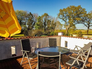 Eine Terrasse im Freien mit Tisch und Stühlen, einem gelb-gestreiften Sonnenschirm und Blick auf Bäume und Felder.