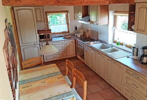 Una cocina con gabinetes blancos, una mesa de comedor con sillas, una lámpara colgante y una ventana con vistas al verde exterior.