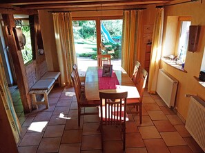 Un coin repas avec une table en bois, des chaises et un banc. Il y a une fenêtre avec des rideaux, un radiateur et une vue sur un jardin avec un toboggan.