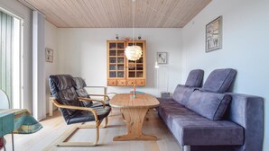 Een gezellige woonkamer met een houten plafond, een ronde houten tafel, een zwarte leren fauteuil, een paarse bank en een kast met glazen deuren.