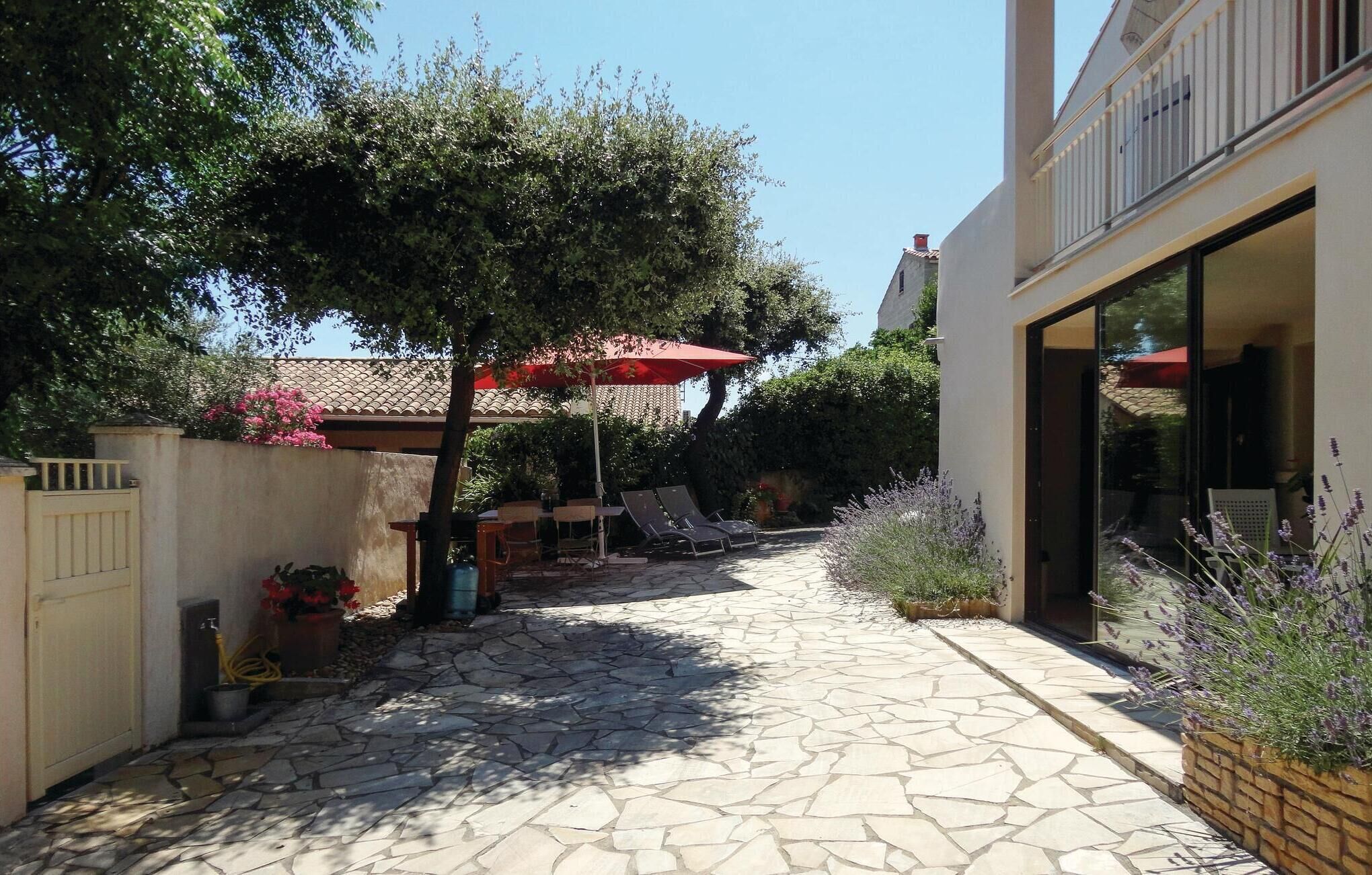 A stone-paved patio with outdoor furniture and a red umbrella, surrounded by greenery and a building with large windows.