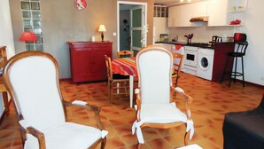 A living room with two wooden chairs, a dining table with a colorful tablecloth, a red cabinet, and a kitchen area with a stove, washing machine, and various kitchen utensils.