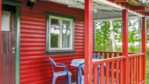Een rode houten chalet met een veranda, een raam en twee blauwe plastic stoelen.