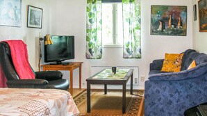 Een woonkamer met een televisie, een bank, een stoel, een bijzettafeltje en een raam met gordijnen.