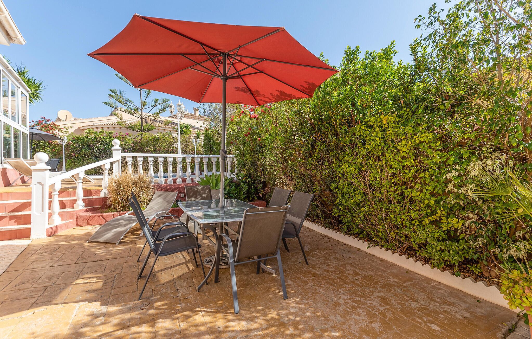Eine Terrasse im Freien mit einem Glastisch und Stühlen, einem roten Sonnenschirm und einer weißen Brüstung.