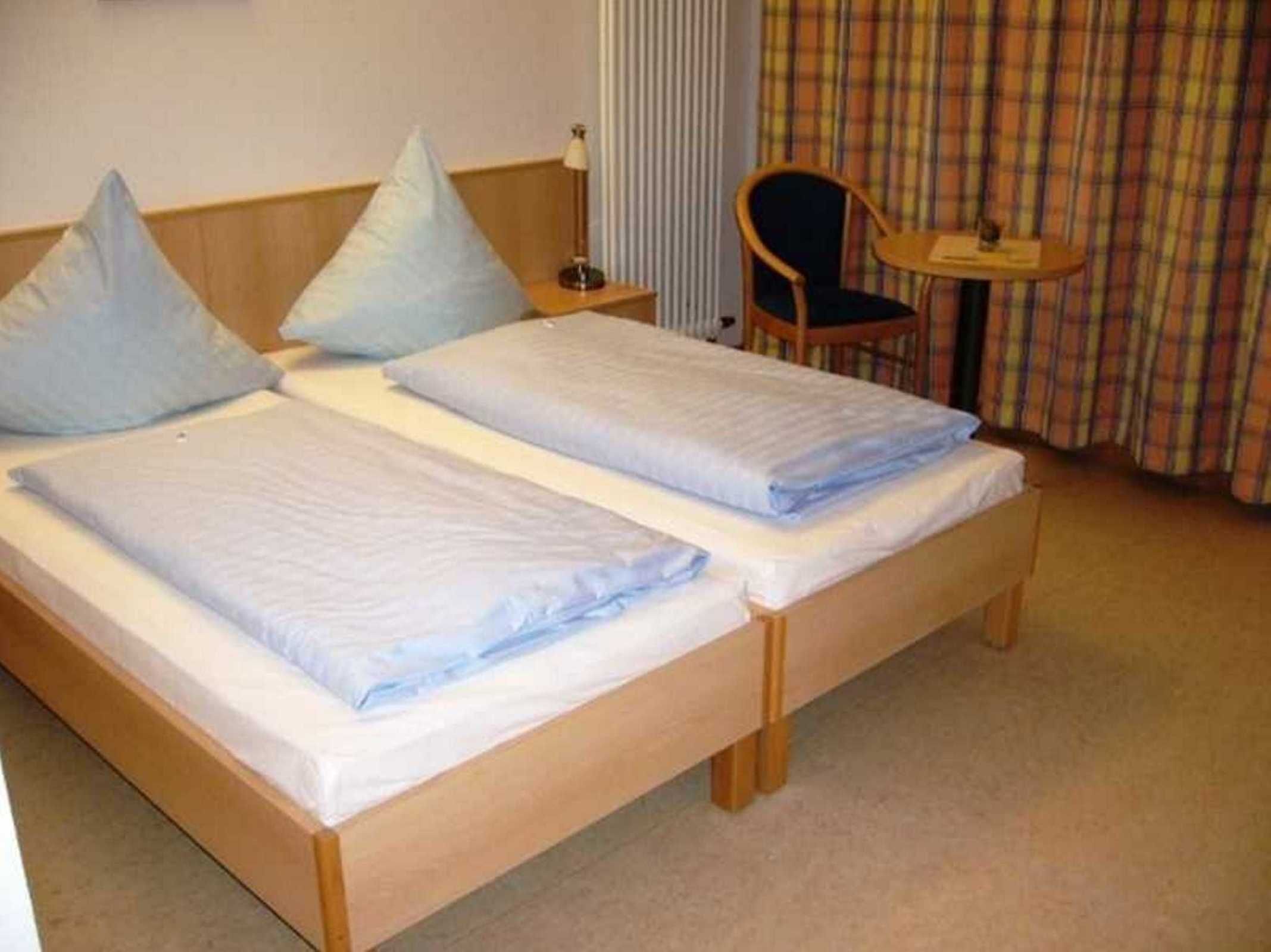 A double bed with a wooden frame, two pillows, a bedside table with a lamp, and a chair with a table.