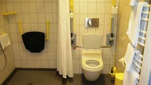 A small, tiled bathroom with a toilet, a hand dryer, a trash bin, and a shower curtain. - Double Room Disability Access - Ferienpark Plauer See (Alt Schwerin)