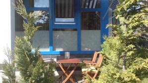 Ein modernes Gebäude mit blauen und weißen Außenwänden, einer überdachten Terrasse mit Holzmöbeln und umliegender Bepflanzung.