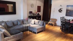 Ein Wohnzimmer mit Ecksofa, einem kleinen runden Tisch, einem Esstisch mit Stühlen, einer Wanduhr und einem Gemälde.