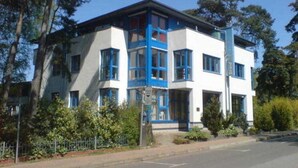 Ein zweistöckiges Gebäude mit weißer Fassade und blauen Akzenten, umgeben von Bäumen und einem Zaun.