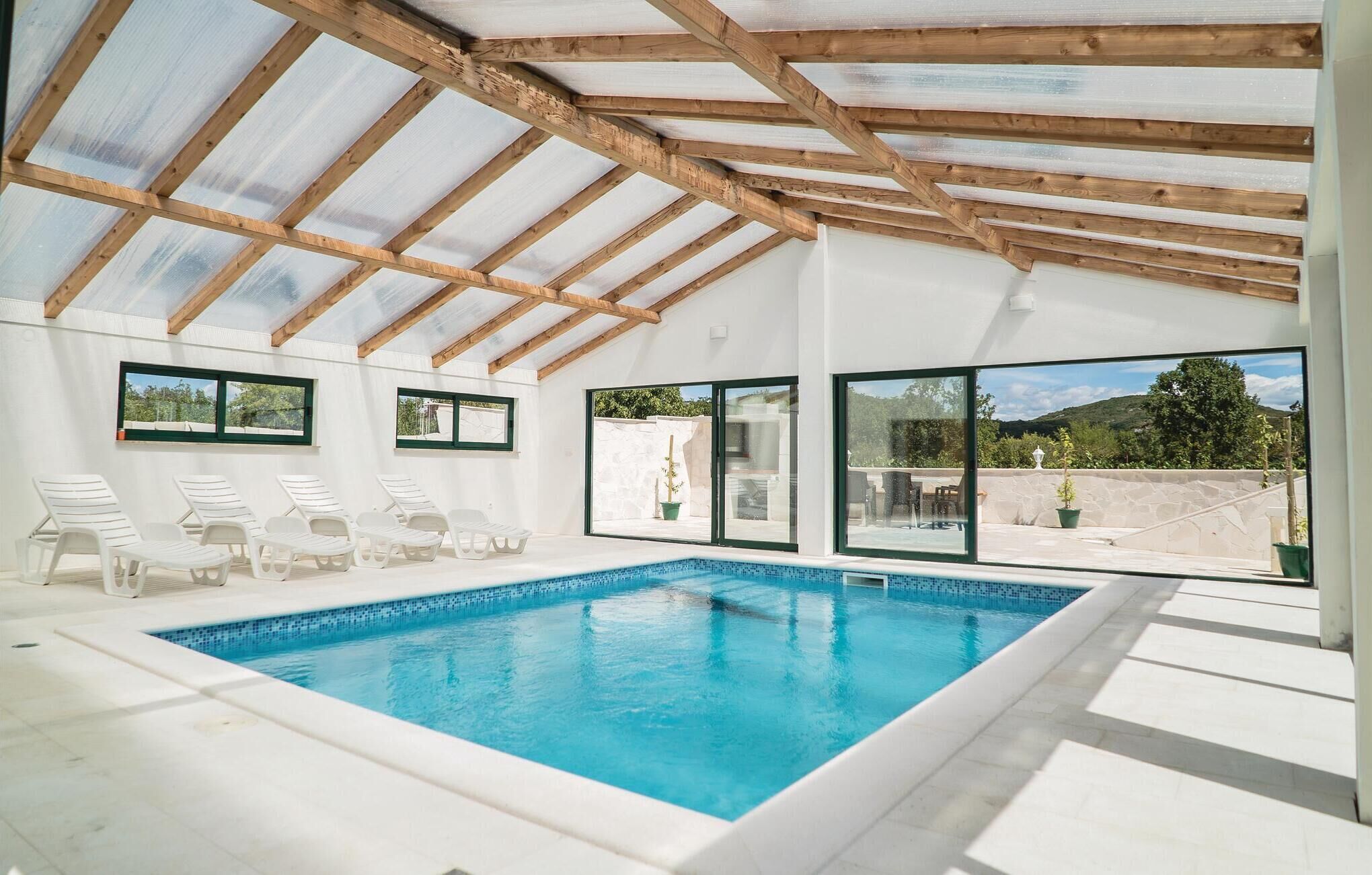 An indoor swimming pool with clear views of the outdoors, wooden beams on the ceiling, and white lounge chairs.