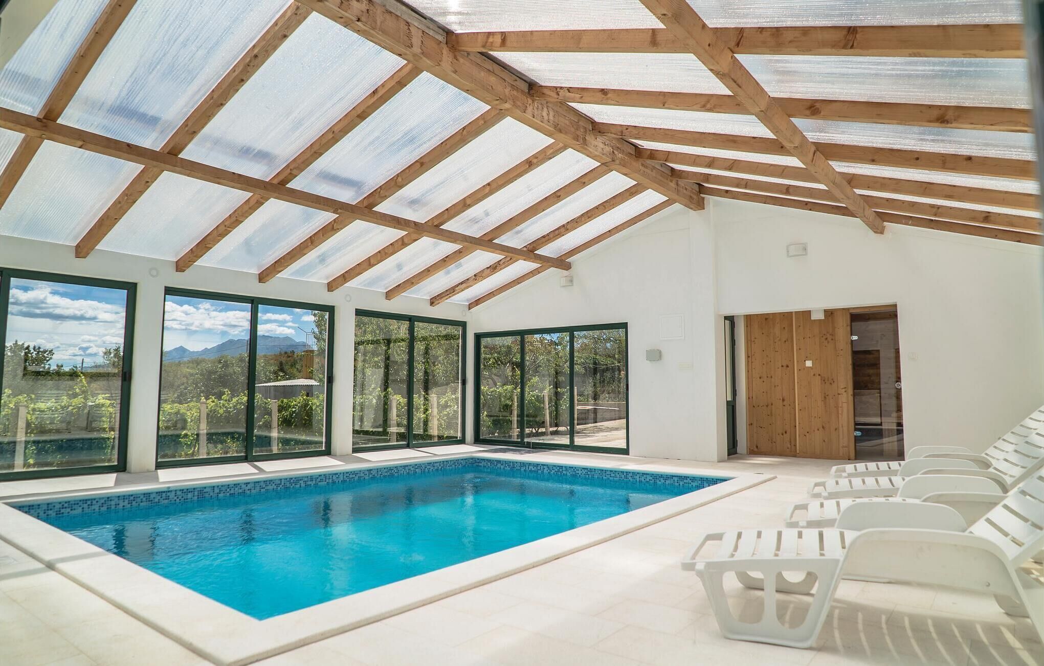 A swimming pool with clear blue water, surrounded by white lounge chairs, large glass doors, and a wooden ceiling structure.