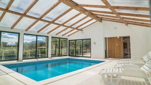 A swimming pool with clear blue water, surrounded by white lounge chairs, large glass doors, and a wooden ceiling structure.