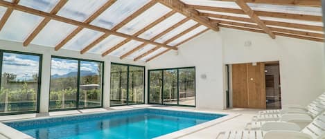 A swimming pool with clear blue water, surrounded by white lounge chairs, large glass doors, and a wooden ceiling structure.