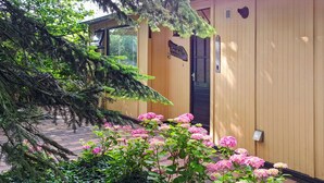 Ein hölzernes Gebäude mit einer schwarzen Tür, umgeben von rosa Hortensienbüschen und einem großen Baum.