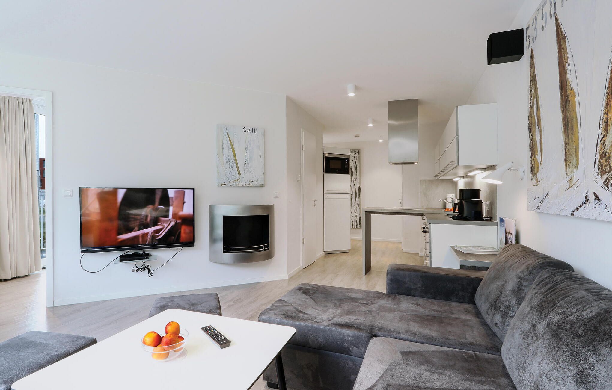 Een moderne woonkamer met een grijze bank, een witte salontafel met fruit en een afstandsbediening, een flatscreen-tv, een open haard en een keuken met een magnetron, fornuis en kastjes.