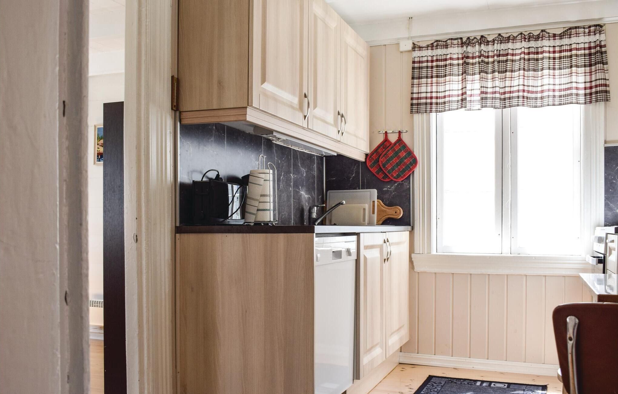 A kitchen with wooden cabinets, a black countertop, a dishwasher, and a window with plaid curtains.