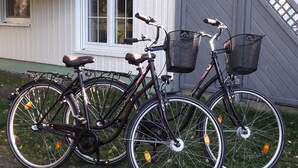 Deux vélos sont garés sur une surface pavée, bordée d’herbe, à proximité d’un bâtiment doté d’une fenêtre et d’une porte.
