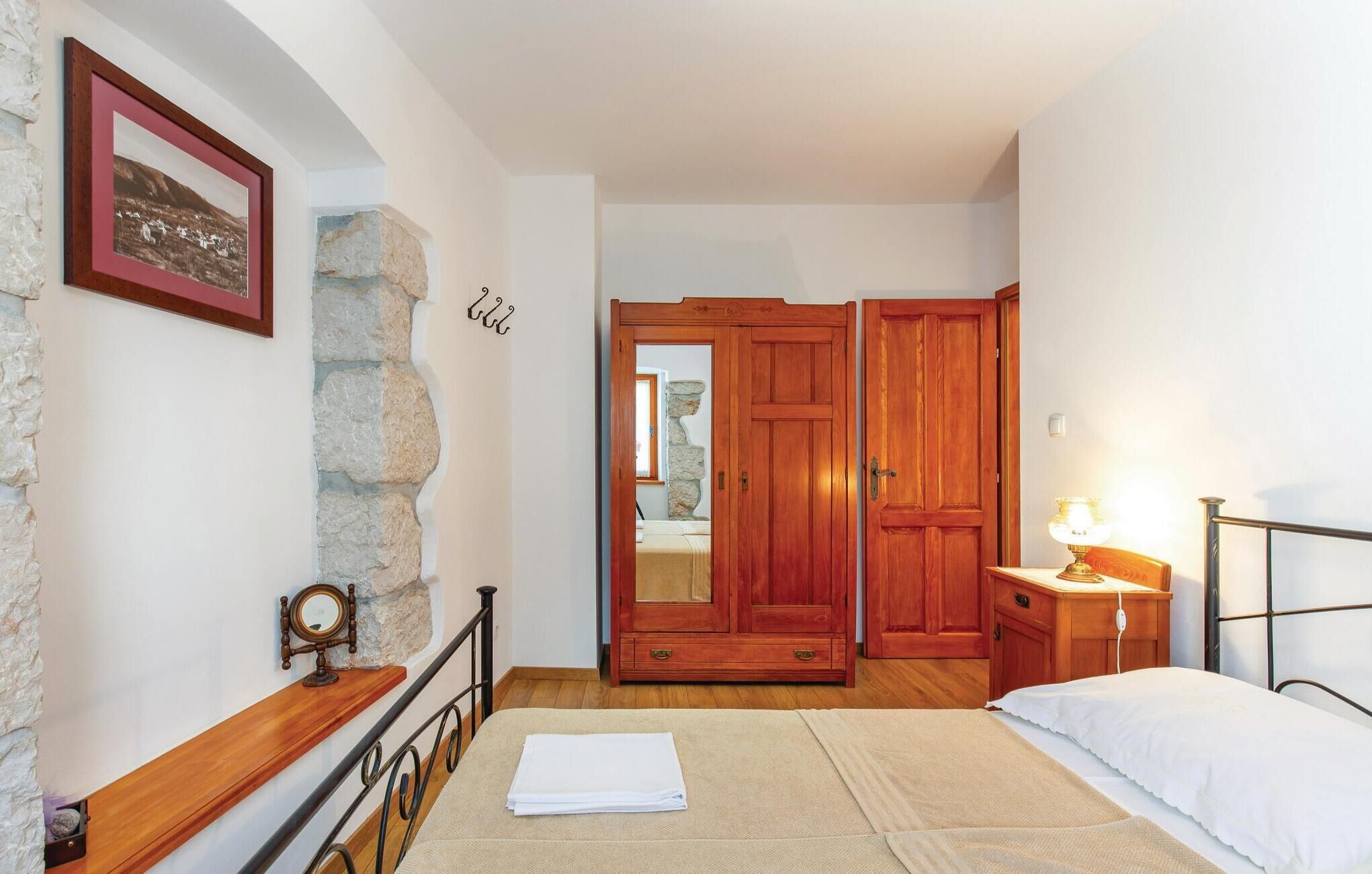 A bedroom with a bed, a nightstand, a wooden wardrobe, a stone wall, and a framed picture on the wall.
