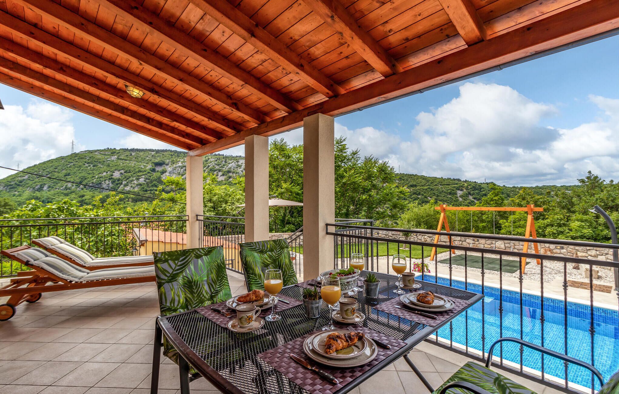 Ein überdachter Essbereich im Freien mit Blick auf einen Pool, mit Tischen, die für ein Frühstück gedeckt sind, darunter Croissants und Orangensaft, sowie Liegestühle.