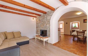 Una sala de estar con pared de piedra, vigas de madera, un sofá, un televisor y una zona de comedor con una mesa y sillas.