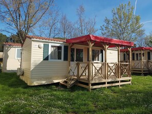 A row of small, single-story cabins with a red roof and wooden deck, surrounded by grass and trees. - Victoria Mobilehome Bi Village (Fazana)