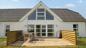 Un bâtiment blanc doté d’un toit à pignon et de grandes fenêtres. On y trouve une terrasse avec un banc et des chaises en bois.