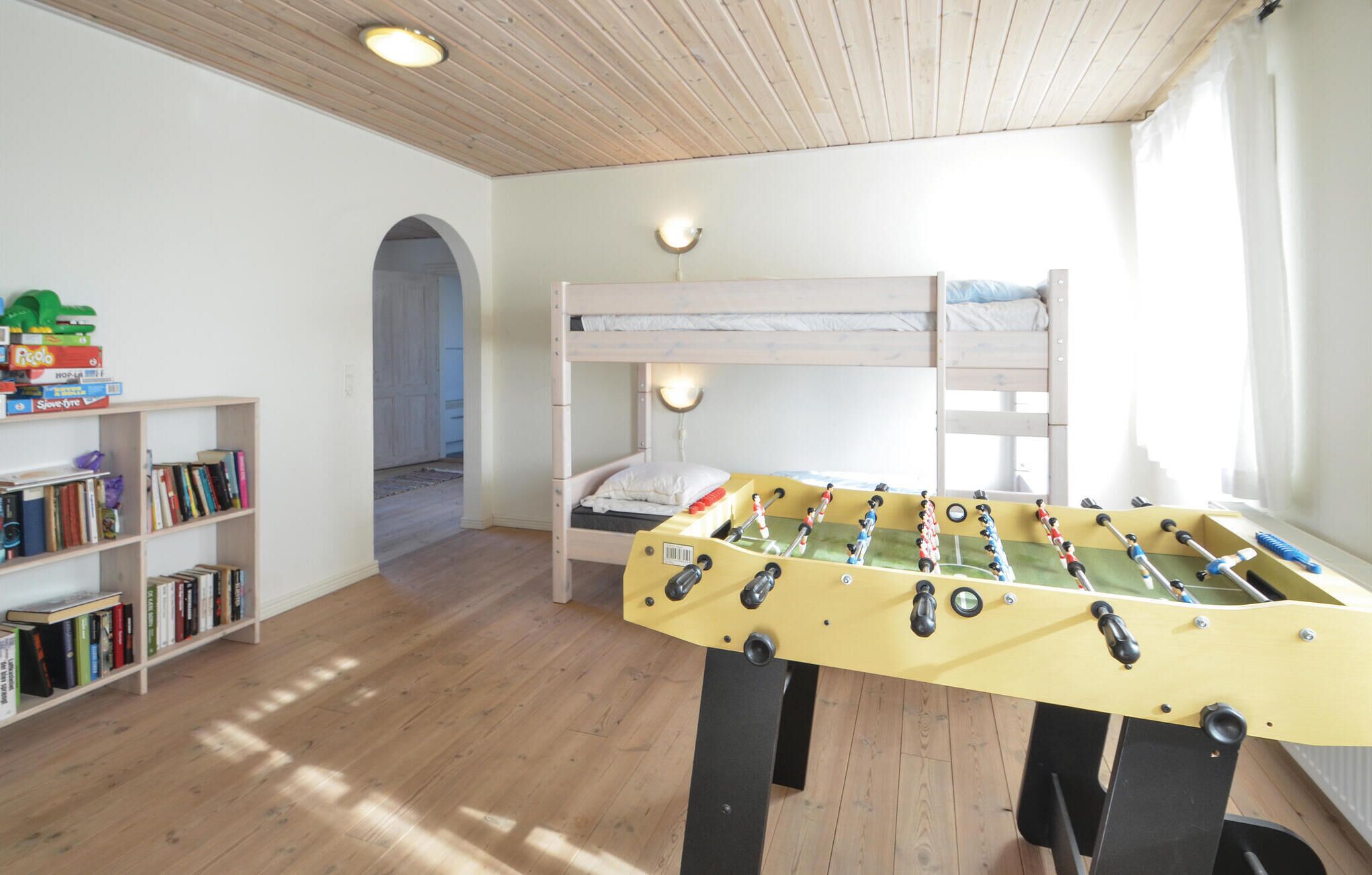 Een kamer met een houten plafond, een tafelvoetbaltafel, een stapelbed, een boekenplank en een deur naar een andere kamer.