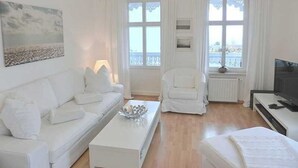 Un soggiorno arredato con mobili bianchi, che include un divano, una poltrona e un tavolino da caffè. Si vede una televisione e una finestra con una vista panoramica.