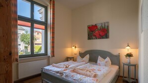 A bedroom with a bed, two bedside tables, a window with curtains, a radiator, and a painting on the wall.