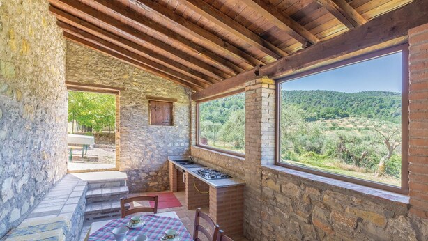 A rustic stone room with a wooden ceiling, a table with chairs, and a view of a forested landscape through large windows. - Amazing home in Magione (PG) (Magione (PG))