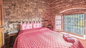 A bedroom with a stone wall, a bed with a pink quilt, a wooden nightstand, a small table with a lamp, and a window with a view of trees. - Amazing home in Magione (PG) (Magione (PG))