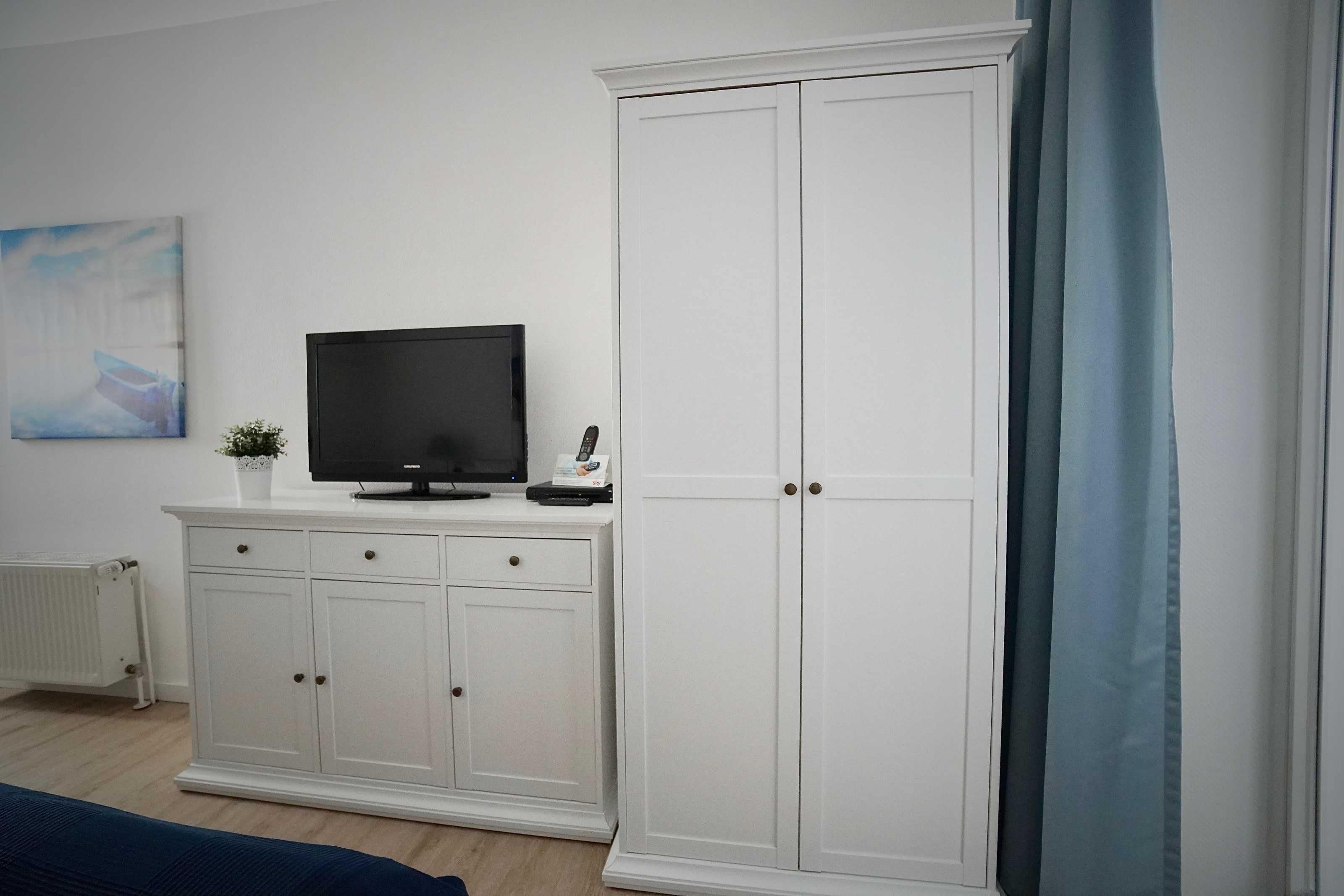 Ein Zimmer mit einem weißen Kleiderschrank, einem Fernseher auf einem Schrank, einem Heizkörper und einem blauen Vorhang.