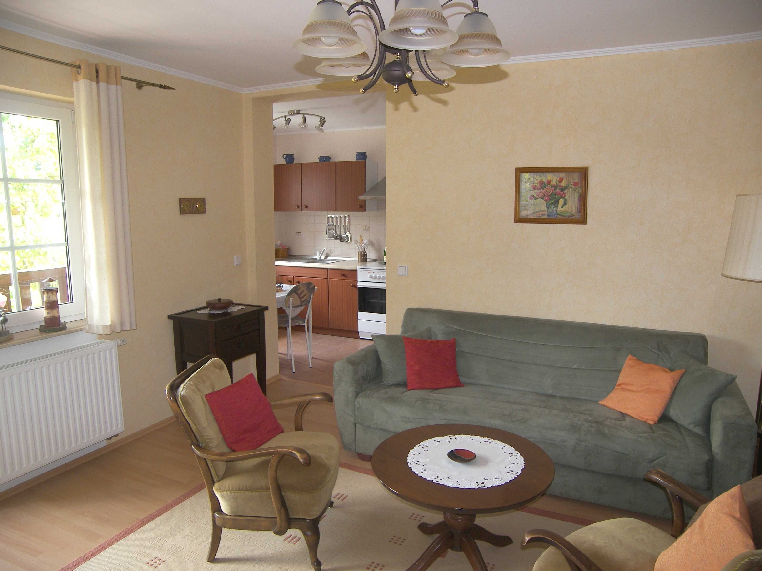 Un salon avec un canapé vert, deux fauteuils, une petite table ronde et, au fond, une cuisine.