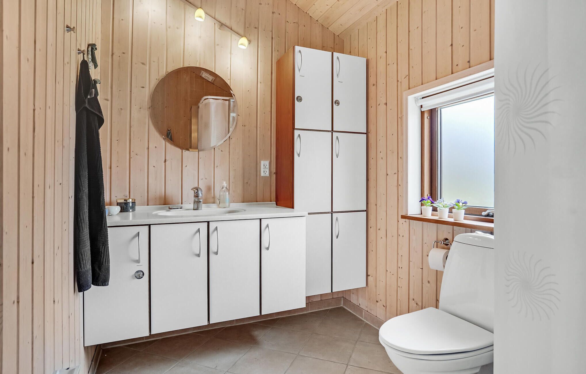A compact bathroom with wooden walls, a white sink, cabinets, a toilet, and a window with plants.