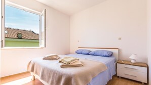 Ein Schlafzimmer mit Bett, Nachttisch, Lampe und einem Fenster mit Blick auf ein Gebäude.