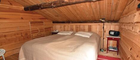 A wooden cabin room with a bed, a small table, a radio, and a chair.