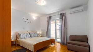 Een slaapkamer met een bed, een stoel, een nachtkastje, een lamp, een raam met gordijnen en een wandairconditioner.